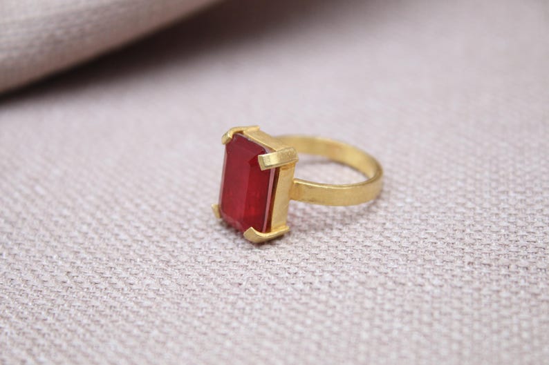 Peut inclure: Une bague dor&eacute;e avec une pierre pr&eacute;cieuse rectangulaire rouge fonc&eacute;. Le corps et le sertissage de la bague ont une finition mate. La pierre est sertie dans un sertissage &agrave; quatre griffes, sur une surface textur&eacute;e et claire.