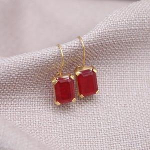 May include: A pair of gold-coloured earrings featuring rectangular, deep red gemstones. The earrings are set in gold-coloured metal and hang from gold-coloured hooks. The background is a light grey, textured fabric.