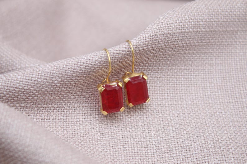 May include: A pair of gold-coloured earrings featuring rectangular, deep red gemstones. The earrings are set in gold-coloured metal and hang from gold-coloured hooks. The background is a light grey, textured fabric.