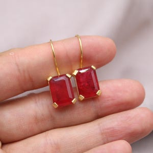 May include: Gold-tone earrings with large, rectangular, deep red gemstones. The gemstones are set in gold bezels and suspended from delicate gold hooks. The earrings are displayed against a neutral background.