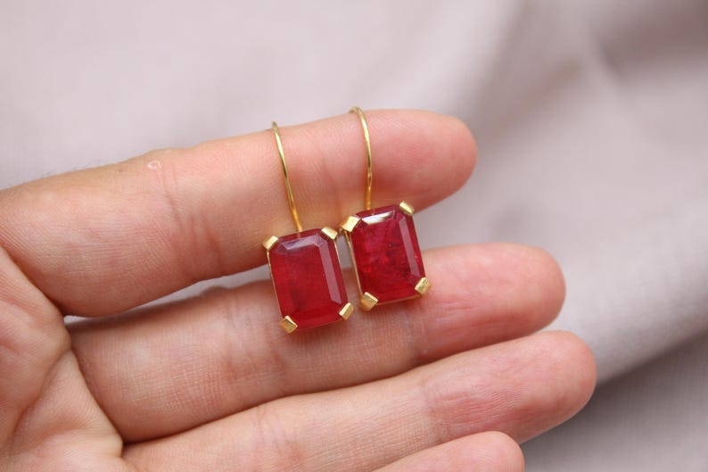 May include: Gold-tone earrings with large, rectangular, deep red gemstones. The gemstones are set in gold bezels and suspended from delicate gold hooks. The earrings are displayed against a neutral background.