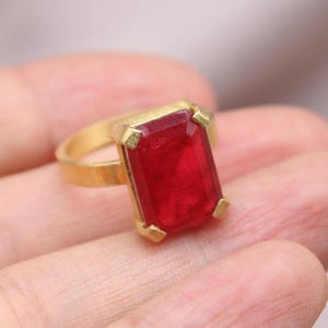 May include: A gold-toned ring featuring a large, rectangular, deep red gemstone. The gemstone is set in a gold bezel with four corner prongs. The ring's band is a simple, polished gold.