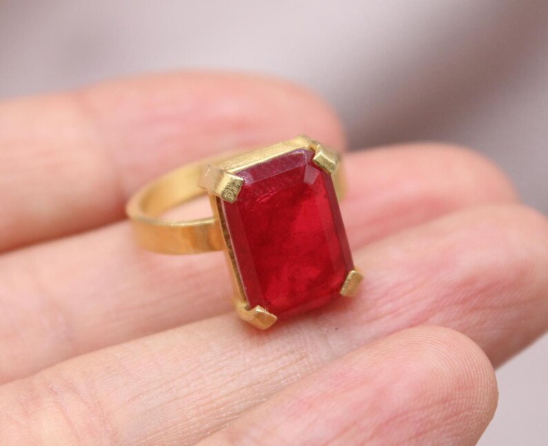 Peut inclure: Une bague dor&eacute;e orn&eacute;e d'une grande pierre pr&eacute;cieuse rouge fonc&eacute; de forme rectangulaire. La pierre est sertie dans une lunette dor&eacute;e avec quatre griffes d'angle. L'anneau est en or poli.