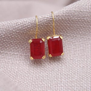 May include: Gold-coloured earrings featuring rectangular, deep red gemstones. The earrings are set in gold and hang from fine gold hooks. The gemstones have a faceted appearance, reflecting light. The earrings rest on a textured, neutral-coloured fabric.