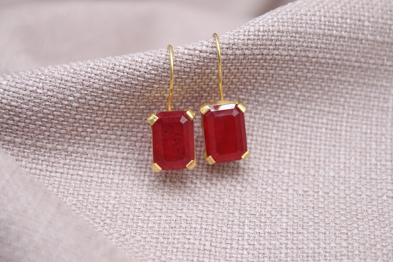 May include: Gold-coloured earrings featuring rectangular, deep red gemstones. The earrings are set in gold and hang from fine gold hooks. The gemstones have a faceted appearance, reflecting light. The earrings rest on a textured, neutral-coloured fabric.