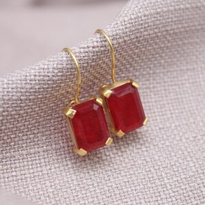 May include: A pair of gold-coloured earrings featuring rectangular, deep red gemstones. The earrings are set in a gold frame and hang from gold-coloured hooks. The earrings are displayed on a textured, light grey fabric.