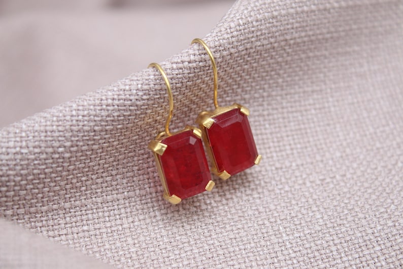 May include: A pair of gold-coloured earrings featuring rectangular, deep red gemstones. The earrings are set in a gold frame and hang from gold-coloured hooks. The earrings are displayed on a textured, light grey fabric.