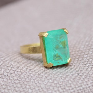 May include: A gold ring featuring a large, rectangular, emerald-green gemstone. The ring has a simple band and gold prongs at each corner of the stone. The gemstone has a slightly textured appearance.