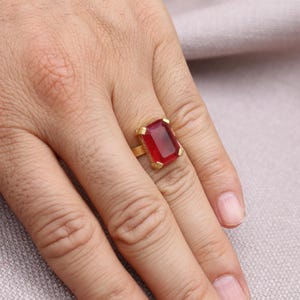 Peut inclure: Une bague en or orn&eacute;e d'une grande pierre rouge rectangulaire. La bague est port&eacute;e au doigt, mettant en valeur la couleur vibrante de la pierre. Le design de la bague est simple, soulignant la beaut&eacute; de la pierre.