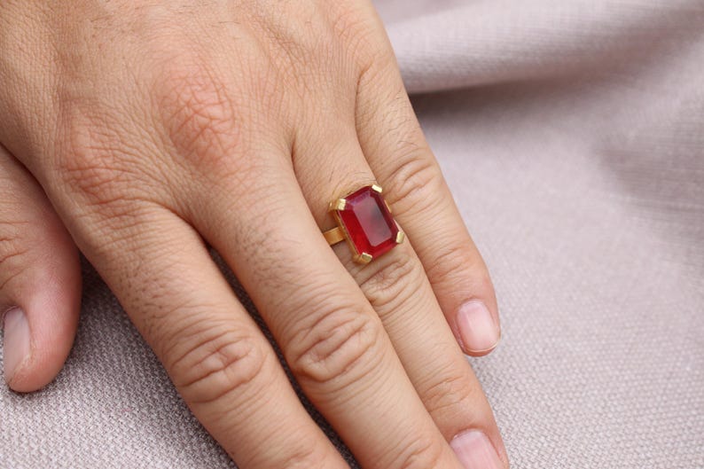 Peut inclure: Une bague en or orn&eacute;e d'une grande pierre rouge rectangulaire. La bague est port&eacute;e au doigt, mettant en valeur la couleur vibrante de la pierre. Le design de la bague est simple, soulignant la beaut&eacute; de la pierre.