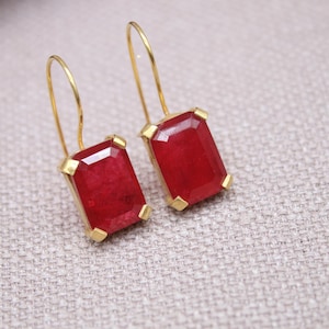 May include: A pair of gold earrings with rectangular, deep red gemstones. The gemstones are set in gold frames and hang from delicate gold wires. The earrings are displayed on a neutral, textured surface.