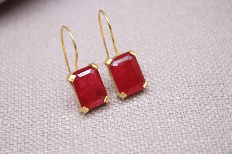 May include: A pair of gold earrings with rectangular, deep red gemstones. The gemstones are set in gold frames and hang from delicate gold wires. The earrings are displayed on a neutral, textured surface.