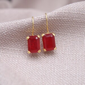 May include: Gold-coloured earrings featuring rectangular, deep red gemstones. The earrings are presented on a textured, light grey fabric. The gemstones are set in gold-coloured metal with a simple hook closure.