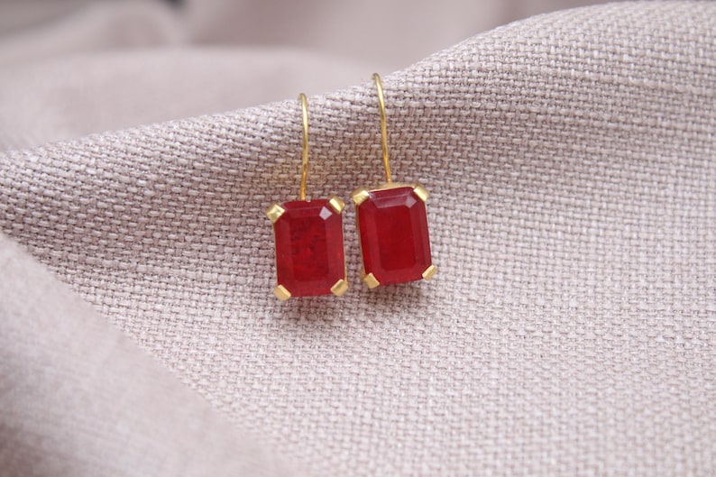 May include: Gold-coloured earrings featuring rectangular, deep red gemstones. The earrings are presented on a textured, light grey fabric. The gemstones are set in gold-coloured metal with a simple hook closure.
