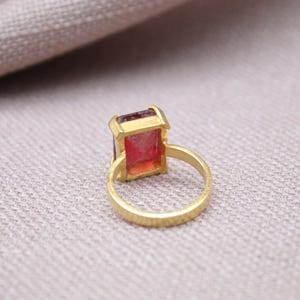 Peut inclure: Une bague dor&eacute;e orn&eacute;e d'une pierre pr&eacute;cieuse rectangulaire rose-rouge. Le corps de la bague pr&eacute;sente une surface textur&eacute;e. La pierre est sertie dans une lunette dor&eacute;e. La bague est pr&eacute;sent&eacute;e sur un tissu de couleur neutre.