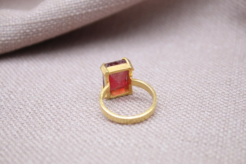 Peut inclure: Une bague dor&eacute;e orn&eacute;e d'une pierre pr&eacute;cieuse rectangulaire rose-rouge. Le corps de la bague pr&eacute;sente une surface textur&eacute;e. La pierre est sertie dans une lunette dor&eacute;e. La bague est pr&eacute;sent&eacute;e sur un tissu de couleur neutre.