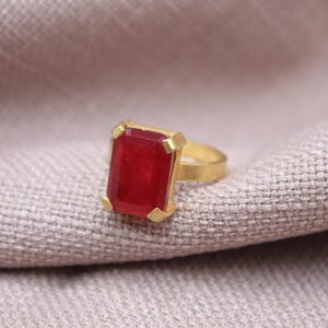 Peut inclure: Une bague dor&eacute;e orn&eacute;e d'une grande pierre pr&eacute;cieuse rouge fonc&eacute; de forme rectangulaire. La pierre est sertie dans une lunette dor&eacute;e. La bague est pr&eacute;sent&eacute;e sur un tissu de couleur neutre, mettant en valeur le design et la couleur du bijou.