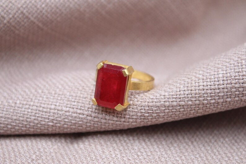 Peut inclure: Une bague dor&eacute;e orn&eacute;e d'une grande pierre pr&eacute;cieuse rouge fonc&eacute; de forme rectangulaire. La pierre est sertie dans une lunette dor&eacute;e. La bague est pr&eacute;sent&eacute;e sur un tissu de couleur neutre, mettant en valeur le design et la couleur du bijou.