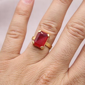 Peut inclure: Une bague en or orn&eacute;e d'une grosse pierre rouge rectangulaire. Le corps de la bague est en or poli et la pierre est sertie de quatre griffes en or. La bague est port&eacute;e sur une main.