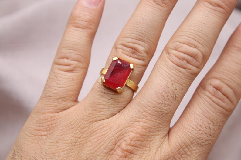 Peut inclure: Une bague en or orn&eacute;e d'une grosse pierre rouge rectangulaire. Le corps de la bague est en or poli et la pierre est sertie de quatre griffes en or. La bague est port&eacute;e sur une main.