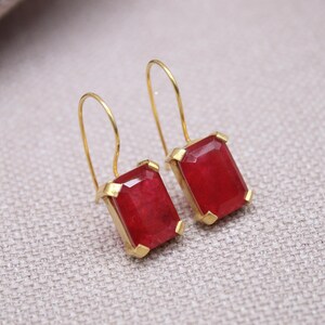 May include: A pair of gold-coloured earrings featuring rectangular, faceted, red gemstones. The earrings have a simple hook design and are set in gold-coloured metal. The gemstones are a vibrant red colour.