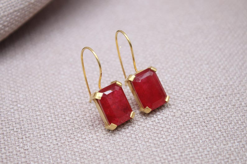 May include: A pair of gold-coloured earrings featuring rectangular, faceted, red gemstones. The earrings have a simple hook design and are set in gold-coloured metal. The gemstones are a vibrant red colour.
