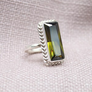 May include: A silver ring with a rectangular, olive-green gemstone. The gemstone is set in a silver frame with a braided design around the edges. The ring is displayed on a textured, light pink surface.