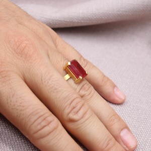 Peut inclure: Une bague en or orn&eacute;e d'une grande pierre rouge rectangulaire. La bague est port&eacute;e au doigt, la main reposant sur une surface de couleur neutre. Le design de la bague est simple, mettant en valeur la couleur vive de la pierre.