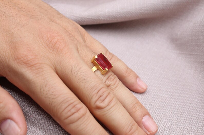 Peut inclure: Une bague en or orn&eacute;e d'une grande pierre rouge rectangulaire. La bague est port&eacute;e au doigt, la main reposant sur une surface de couleur neutre. Le design de la bague est simple, mettant en valeur la couleur vive de la pierre.