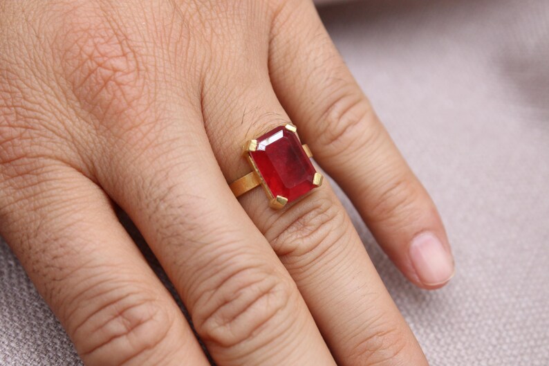 Peut inclure: Une bague en or orn&eacute;e d'une grande pierre pr&eacute;cieuse rouge fonc&eacute; de forme rectangulaire. Le sertissage de la bague est simple et &eacute;l&eacute;gant, la pierre pr&eacute;cieuse &eacute;tant le point central. La bague est port&eacute;e sur une main.