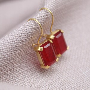 May include: A pair of gold earrings featuring rectangular, deep red gemstones. The earrings are presented on a textured, light grey fabric. The gemstones have a faceted cut, reflecting light.