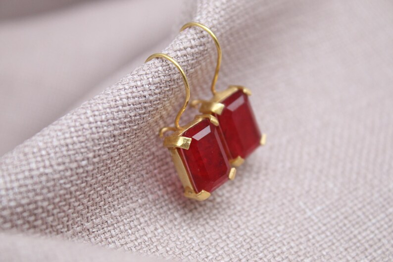 May include: A pair of gold earrings featuring rectangular, deep red gemstones. The earrings are presented on a textured, light grey fabric. The gemstones have a faceted cut, reflecting light.