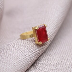 Peut inclure: Une bague en or avec une grosse pierre pr&eacute;cieuse rouge fonc&eacute; de forme rectangulaire. La bague est pos&eacute;e sur un tissu gris clair, soulignant le contraste entre l'or chaud et la pierre rouge vibrante. Le design de la bague est simple mais &eacute;l&eacute;gant.