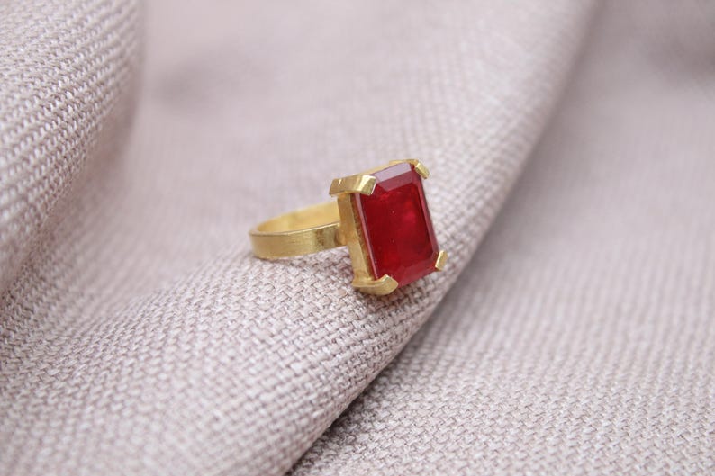 Peut inclure: Une bague en or avec une grosse pierre pr&eacute;cieuse rouge fonc&eacute; de forme rectangulaire. La bague est pos&eacute;e sur un tissu gris clair, soulignant le contraste entre l'or chaud et la pierre rouge vibrante. Le design de la bague est simple mais &eacute;l&eacute;gant.