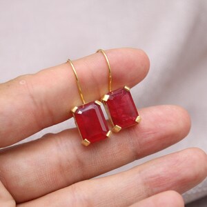 May include: Gold-coloured earrings with rectangular, deep red gemstones. The gemstones are set in a gold frame and suspended from a simple gold hook. The earrings are displayed against a neutral background.