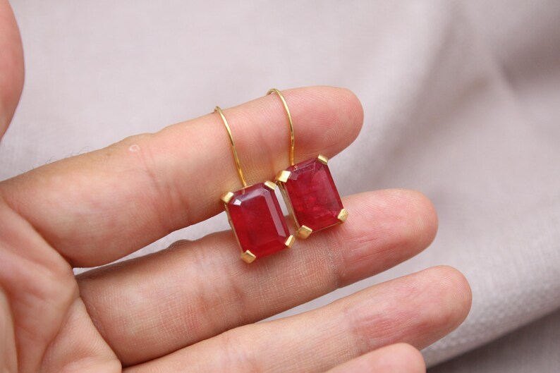 May include: Gold-coloured earrings with rectangular, deep red gemstones. The gemstones are set in a gold frame and suspended from a simple gold hook. The earrings are displayed against a neutral background.