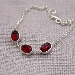 May include: A silver bracelet featuring three oval, deep red gemstones. The gemstones are set in individual silver bezels and connected by a delicate silver chain. The bracelet has a lobster clasp closure.