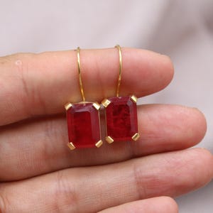 May include: A pair of gold earrings with rectangular, deep red gemstones. The earrings have a simple design with a gold hook and a gold setting around the red stones. The earrings are held in a hand.