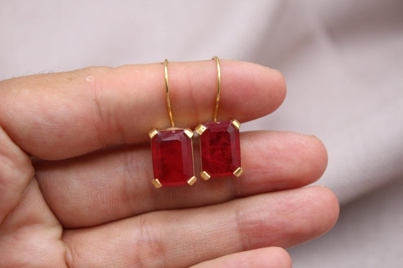 May include: A pair of gold earrings with rectangular, deep red gemstones. The earrings have a simple design with a gold hook and a gold setting around the red stones. The earrings are held in a hand.