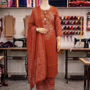 Puede incluir: Un conjunto salwar kameez de color óxido sobre un maniquí. El atuendo presenta una túnica de manga larga, pantalones a juego y una bufanda estampada. La túnica y los pantalones tienen un patrón de puntos blancos. El maniquí está en un estudio de costura.