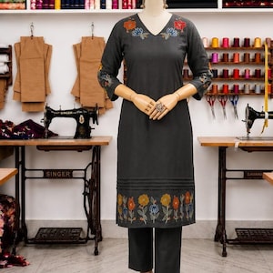 May include: A dark gray embroidered kurta and matching pants are displayed on a mannequin. The kurta features floral embroidery on the neckline, sleeves, and hem. The background shows sewing machines, fabric bolts, and spools of thread.