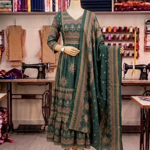 May include: A green dress and matching shawl are displayed on a mannequin. The dress has a V-neck and a detailed gold and brown pattern. The floor-length dress has long sleeves. Sewing machines and fabric bolts are visible in the background.