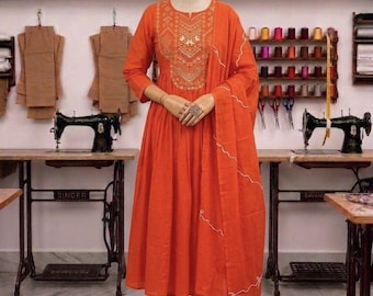 May include: A bright orange long-sleeved dress with a flared skirt and matching trousers. The bodice features intricate gold-colored embroidery around the neckline. A coordinating orange shawl with a thin white wavy line pattern is draped over one arm.
