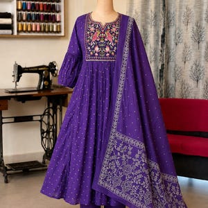 May include: A deep purple long-sleeved dress with a gathered waist and a matching dupatta. The bodice features intricate multi-colored floral and bird embroidery around the neckline. The fabric has a small white repeating pattern throughout.