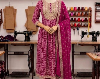 May include: A magenta Indian salwar kameez suit with gold embroidery. The outfit includes a long-sleeved tunic, matching trousers, and a dupatta. The suit is displayed on a mannequin in a sewing studio.