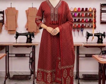 May include: A maroon three-piece outfit featuring a long-sleeved kurta with a V-neckline, intricate embroidery, and mirror accents. The set includes matching slim-fit trousers and a coordinating patterned dupatta draped over the shoulder.