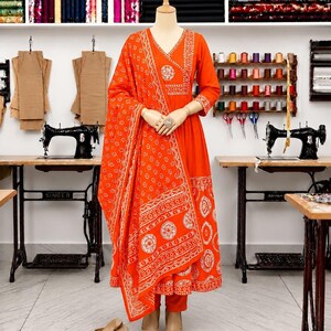 May include: A bright orange long-sleeved dress with a V-neckline and a matching shawl, both featuring white circular and geometric patterns. The dress is paired with orange trousers and beige slip-on shoes, displayed on a mannequin in a studio.
