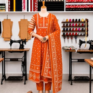 May include: An orange long-sleeved dress with a white geometric and floral pattern, paired with matching orange trousers and a draped shawl. The outfit is displayed on a mannequin in a sewing room with two black sewing machines and thread spools.
