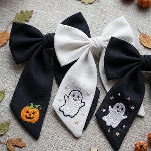 Peut inclure: Trois nœuds pour cheveux en tissu noir et blanc. Un nœud noir présente une citrouille orange brodée. Un nœud blanc présente un fantôme blanc brodé avec de petites étoiles. Un nœud noir présente un fantôme blanc brodé avec des étoiles.