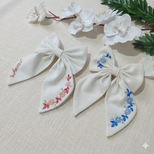 Może przedstawiać: Trzy białawe kokardy do włosów z tkaniny. Dwie kokardy mają kwiatowe hafty w kolorze różowym i niebieskim. Jedna kokarda ma wyhaftowane imię "sirineya" na czerwono. Kokardy ułożone są na jasnej powierzchni.